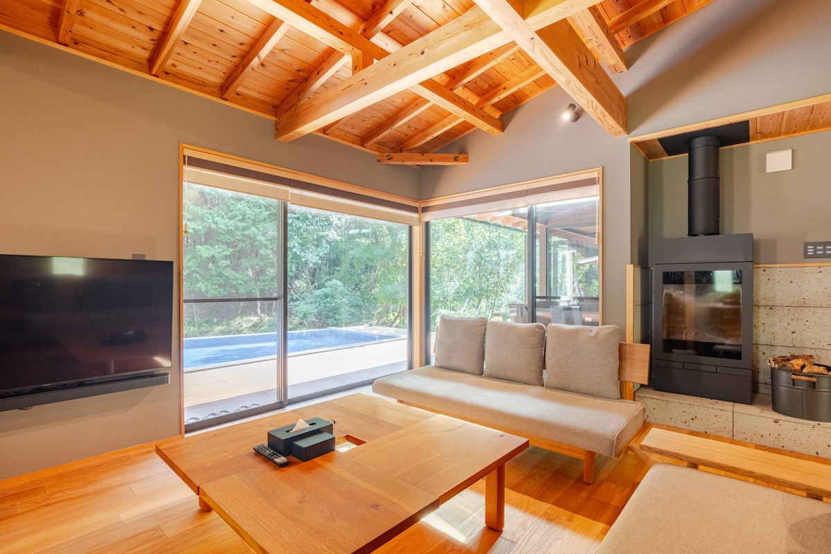 A spacious living area is featured with large glass sliding doors, providing a view of the outdoor pool. A minimalist wooden coffee table sits in the center, accompanied by a low-profile sofa. Natural light fills the room, highlighting the wooden beams of the ceiling.