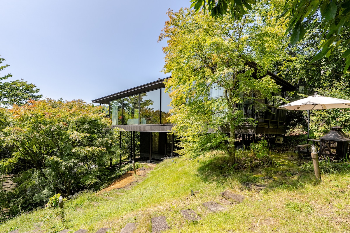The exterior of the house is showcased, featuring large glass panels that reflect the surrounding nature. Lush greenery and trees create a serene environment. A patio area with an umbrella and a stone pathway leads up to the entrance, enhancing the connection with the outdoors.