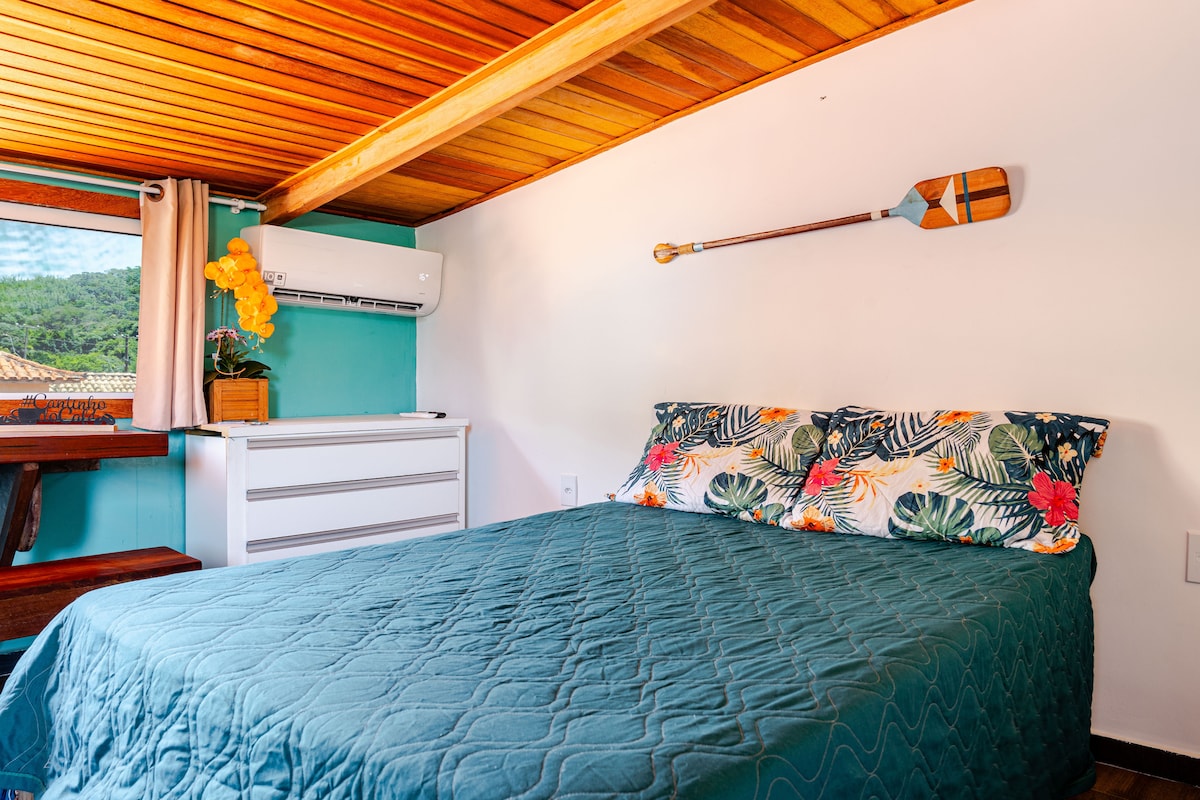 A cozy sleeping area features a spacious bed topped with a floral-patterned blanket. A chest of drawers is located nearby, and a kayak paddle is displayed on the wall above. Natural light enters through a window, complemented by decorative curtains.
