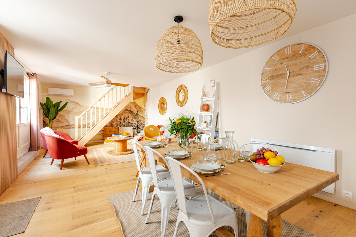 The dining area features a wooden table set for six, surrounded by white chairs. Two stylish pendant lights hang above. A decorative wall clock and potted plant accentuate the space. The staircase is visible in the background, leading to the upper level.