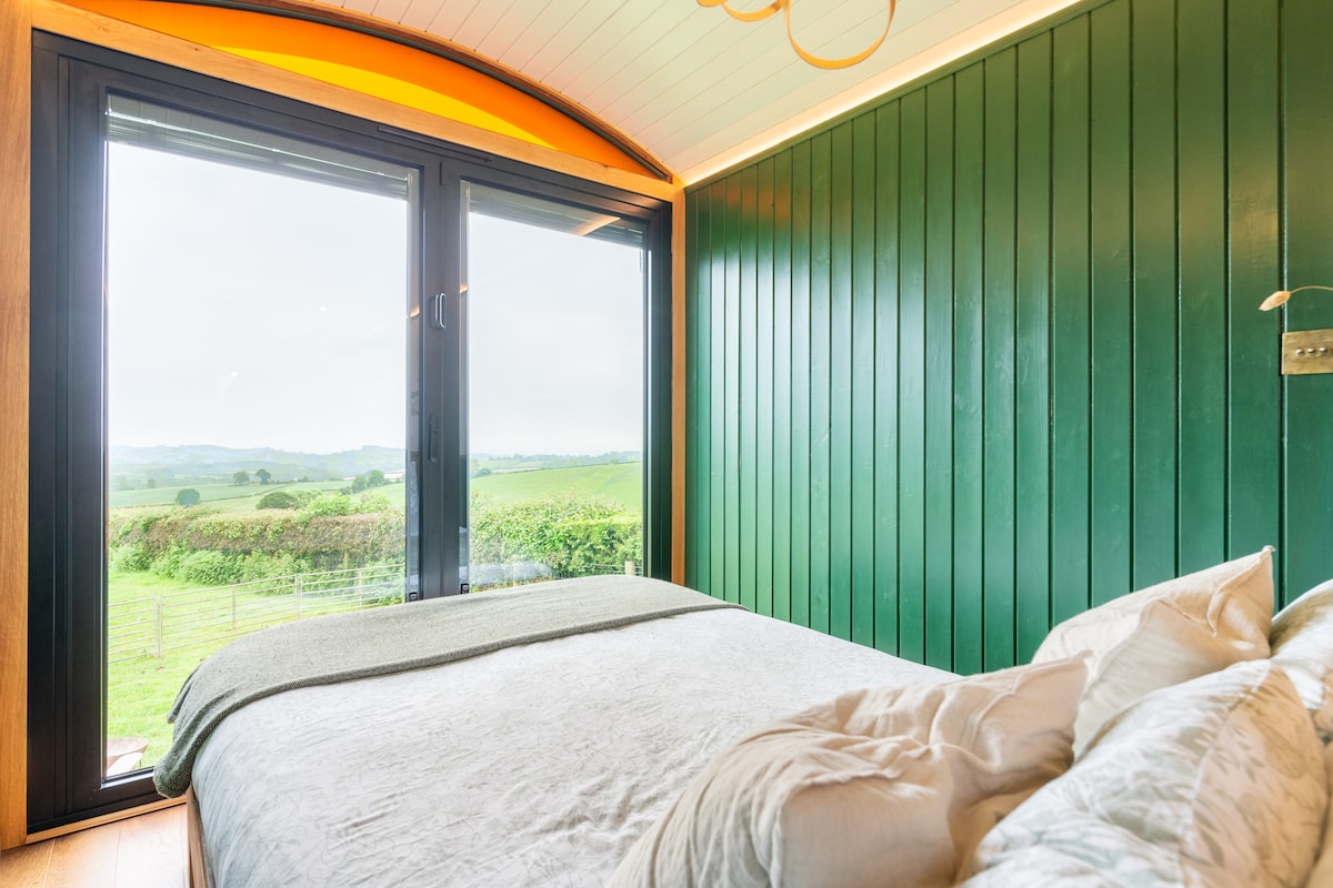 A cozy sleeping area features a spacious bed with soft linens and decorative pillows. Floor-to-ceiling glass doors provide panoramic views of the surrounding countryside. Natural light floods the room, accentuating the rich green wall panels, enhancing the serene atmosphere.