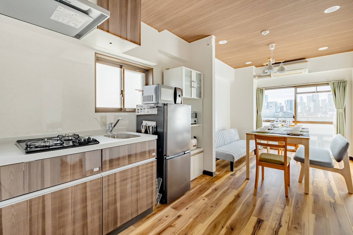 A well-equipped kitchen showcases modern appliances, including a refrigerator, microwave, and gas stove. The dining area features a wooden table with seating for four, complemented by large windows offering a view of the city skyline. Sunlight fills the open, spacious layout.