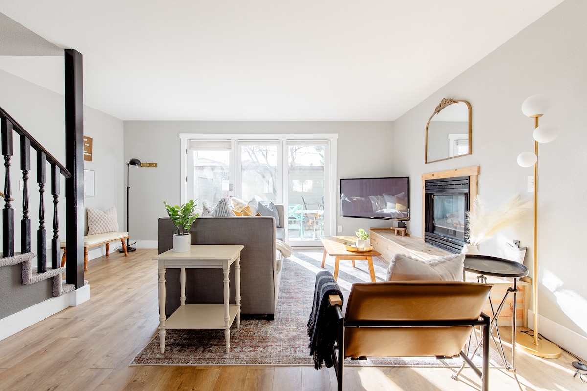 The living area includes an open layout with a large seating arrangement facing a fireplace and a flat-screen TV. Natural light fills the space through large windows, and a coffee table is positioned in front of the couch, enhancing the home's comfort.