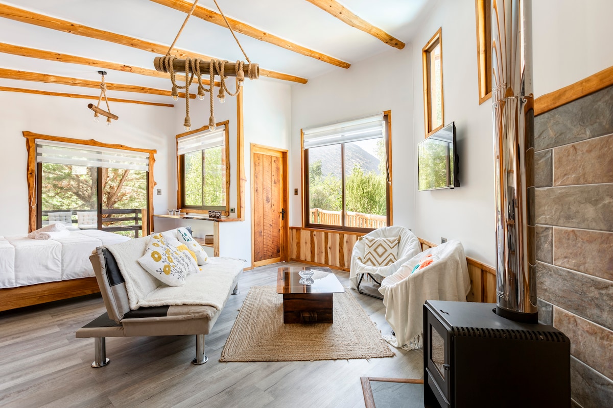 The inviting living area features wooden beams and a cozy sofa adorned with decorative pillows. A flat-screen TV is mounted on the wall across from a rustic coffee table. Natural light filters through large windows, illuminating the warm wood accents and soft textures throughout the space.