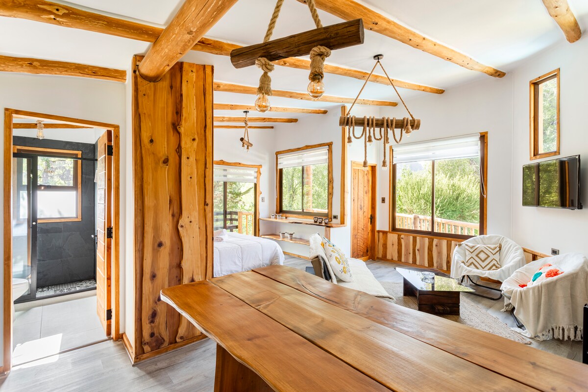 A spacious interior is highlighted by wooden beams and natural textures. Large windows allow light to fill the area, offering views of the lush garden outside. A mix of seating arrangements is visible, along with a dining table made from natural wood.