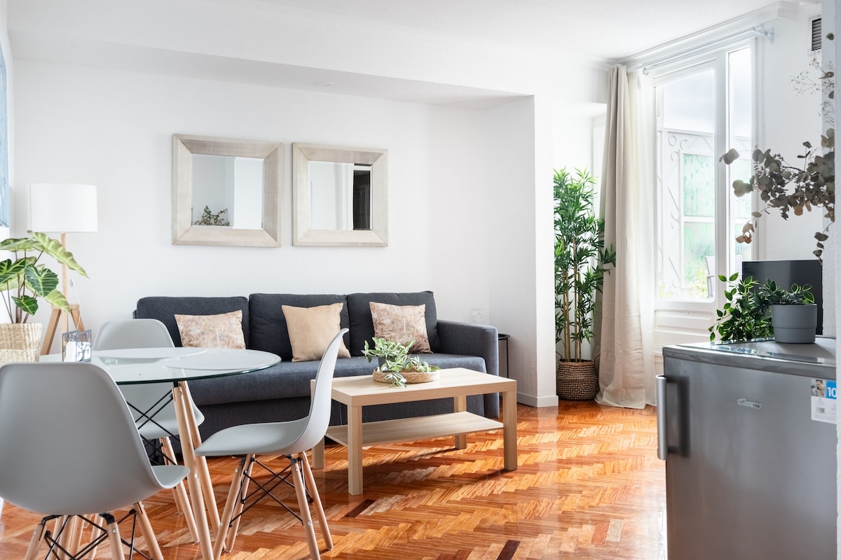A cozy living area features a dark sofa adorned with cushions, positioned across from a circular dining table. Light wood flooring complements the space, while mirrors reflect the natural light from the windows. Decorative plants add a touch of greenery to the inviting atmosphere.