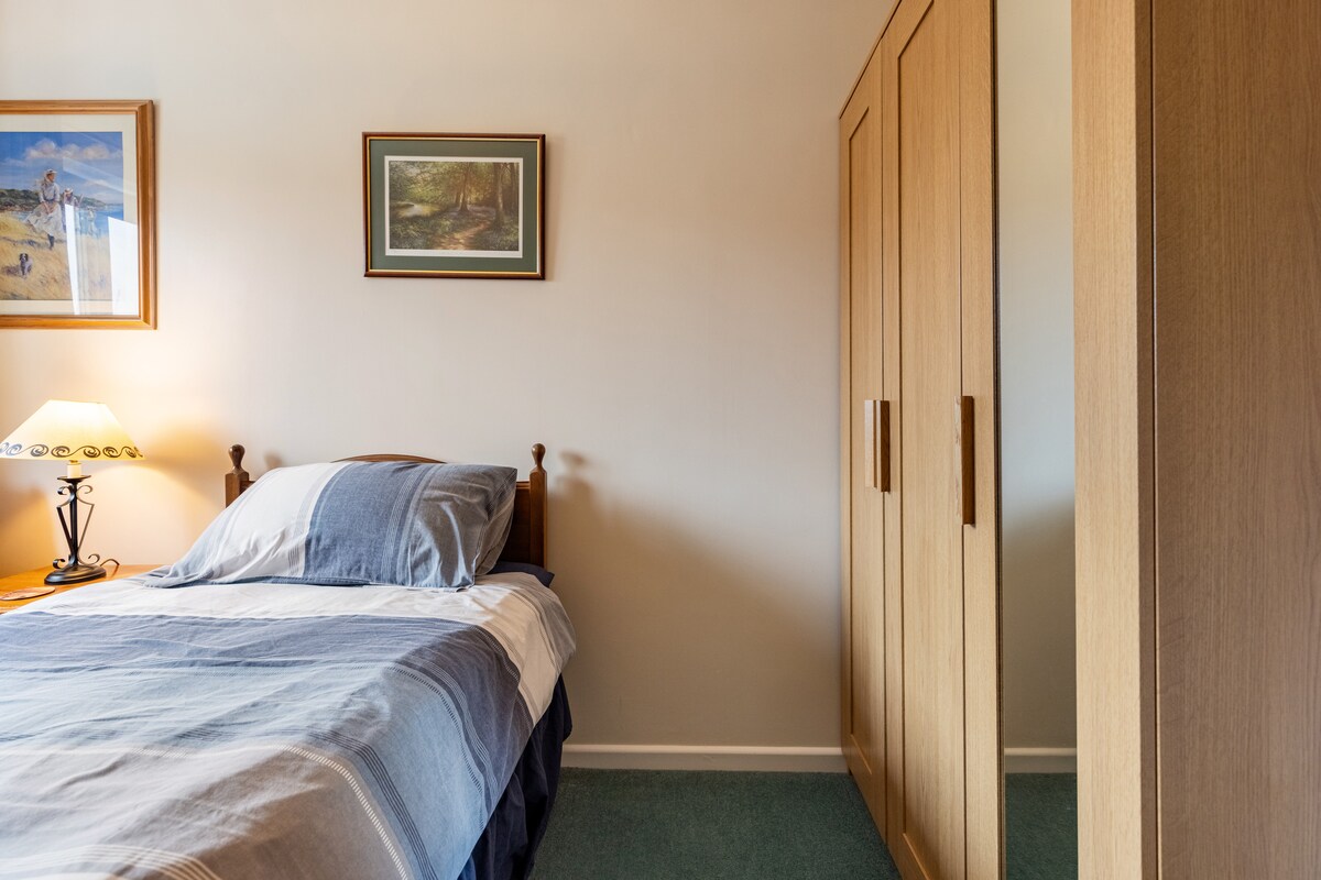 A cozy sleeping area features a single bed with a blue and white striped blanket. A bedside lamp provides soft illumination beside the bed, while a wardrobe with sliding doors is positioned nearby. Artwork adorns the wall, contributing to a tranquil ambiance.