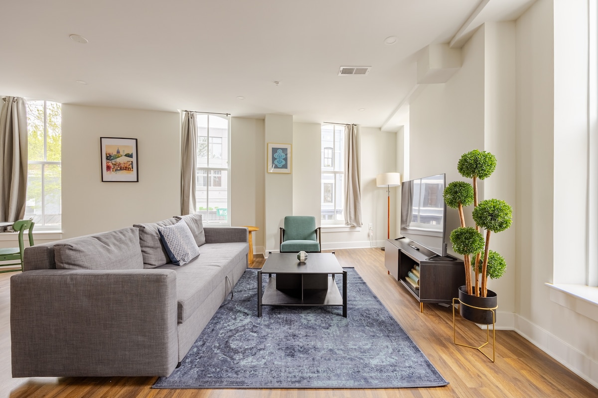 A spacious living area features a large gray sofa positioned beside a coffee table with a decorative plant. A television stands on a dark media unit, and light enters through large windows adorned with sheer curtains. A small green chair complements the modern space.