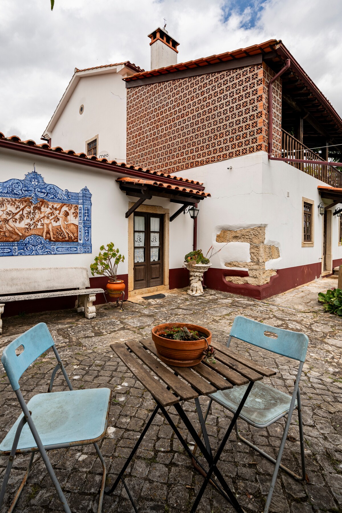 A charming exterior view of the house features a decorative blue tile mural on one wall. A small wooden table with two blue chairs is positioned on a cobblestone patio, accompanied by a terra-cotta pot containing greenery. The structure showcases traditional architectural elements.