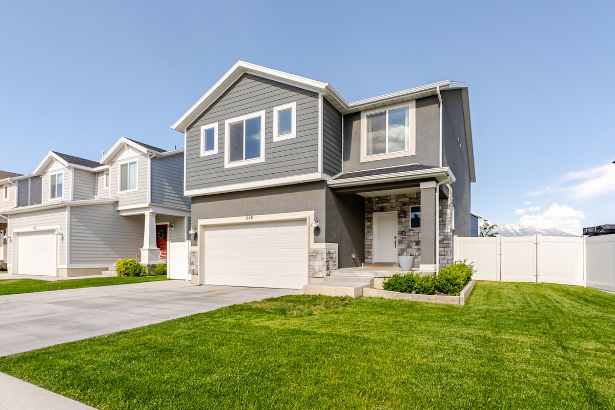 A two-story home is displayed, featuring a modern gray exterior with white accents. The front yard is well-maintained, showcasing lush green grass. A spacious driveway leads to a garage, and neighboring houses provide context to the residential setting.