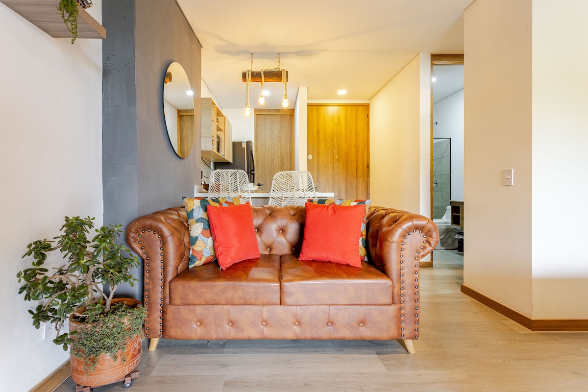 A cozy living area features a brown leather sofa adorned with two vibrant red pillows. A potted plant enhances the space, while light wood flooring and an accent wall in a soft hue create a warm atmosphere. The open layout leads to the kitchen and dining area.