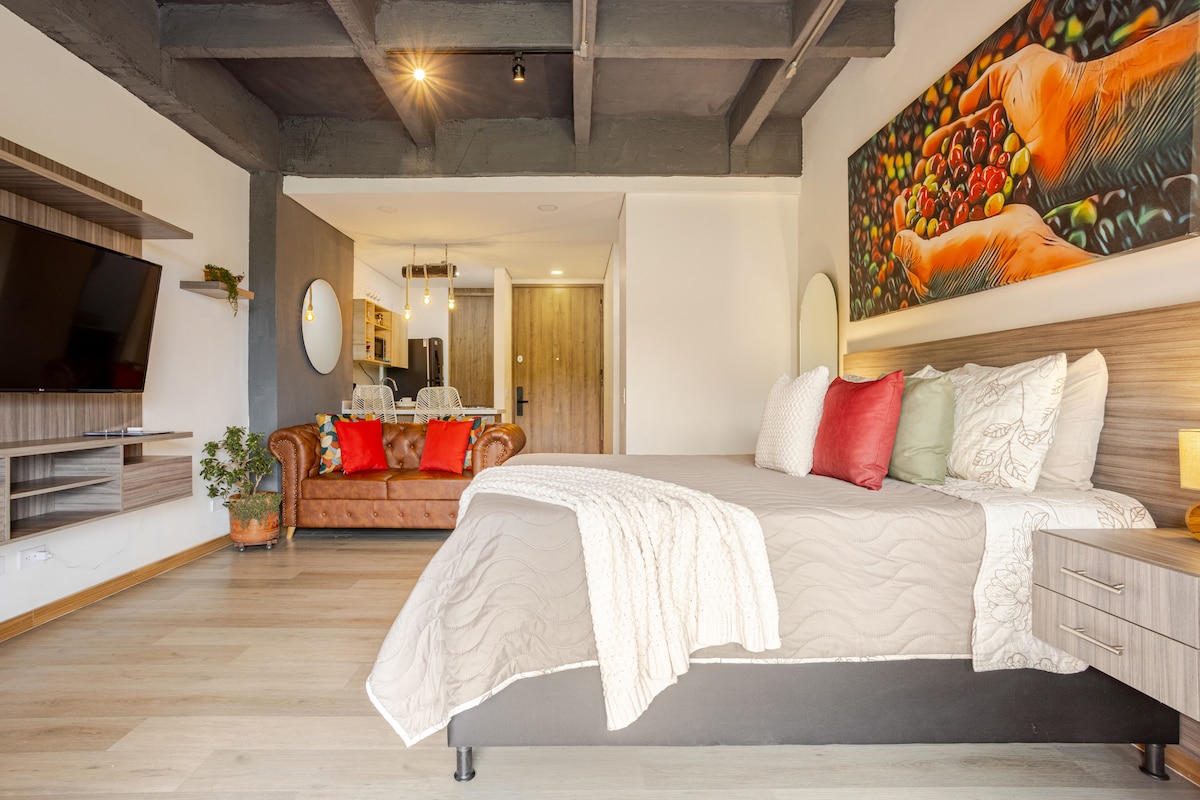 A spacious bedroom features a large bed dressed in neutral tones, complemented by accent pillows in various colors. A wooden media unit houses a TV across from a cozy leather sofa. A vibrant wall art piece depicting coffee beans adds an inviting touch.