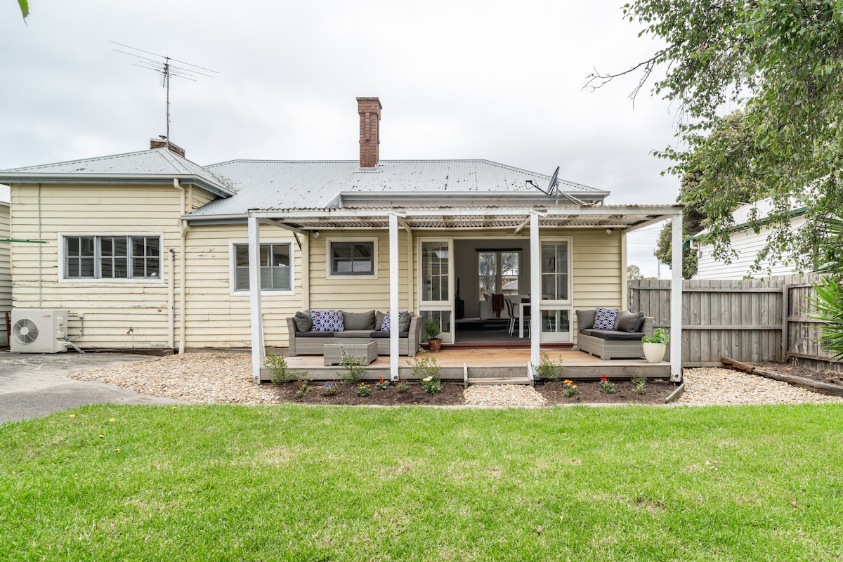The rear view of the home is presented, featuring a covered porch with comfortable outdoor seating. A well-maintained lawn stretches out, bordered by gravel landscaping. The setting offers a sense of tranquility, inviting relaxation in a spacious backyard.