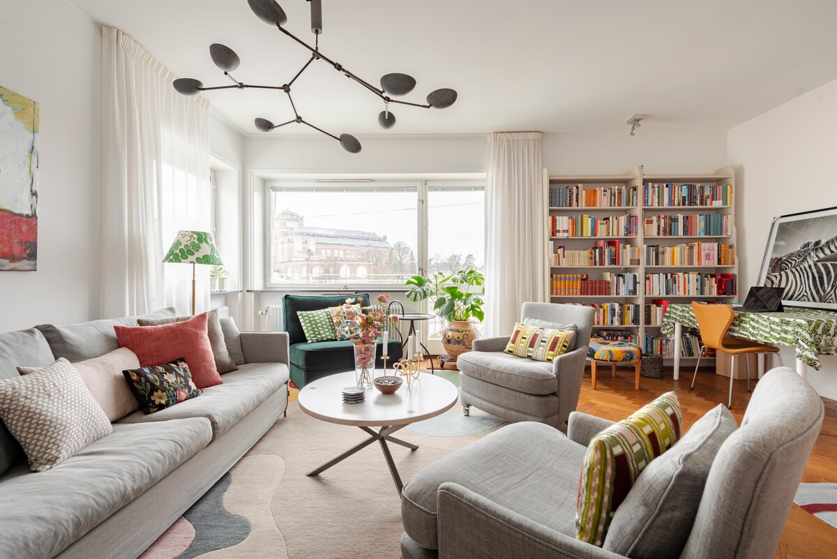 A light-filled living room showcases a comfortable gray sofa accented with patterned pillows. A round coffee table centers the space, surrounded by two armchairs and a bookshelf filled with books. Large windows provide views of the outdoors, with a small plant and a lamp adding warmth.