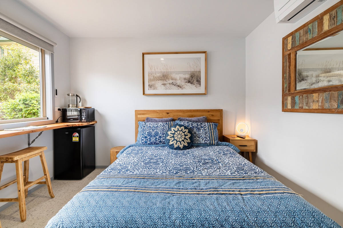 A newly renovated bedroom is presented with a spacious bed adorned in blue bedding. A kitchenette and refrigerator are visible, complemented by a wooden desk and stool. Natural light filters through a window, illuminating a mirror framed in rustic wood on the opposite wall.