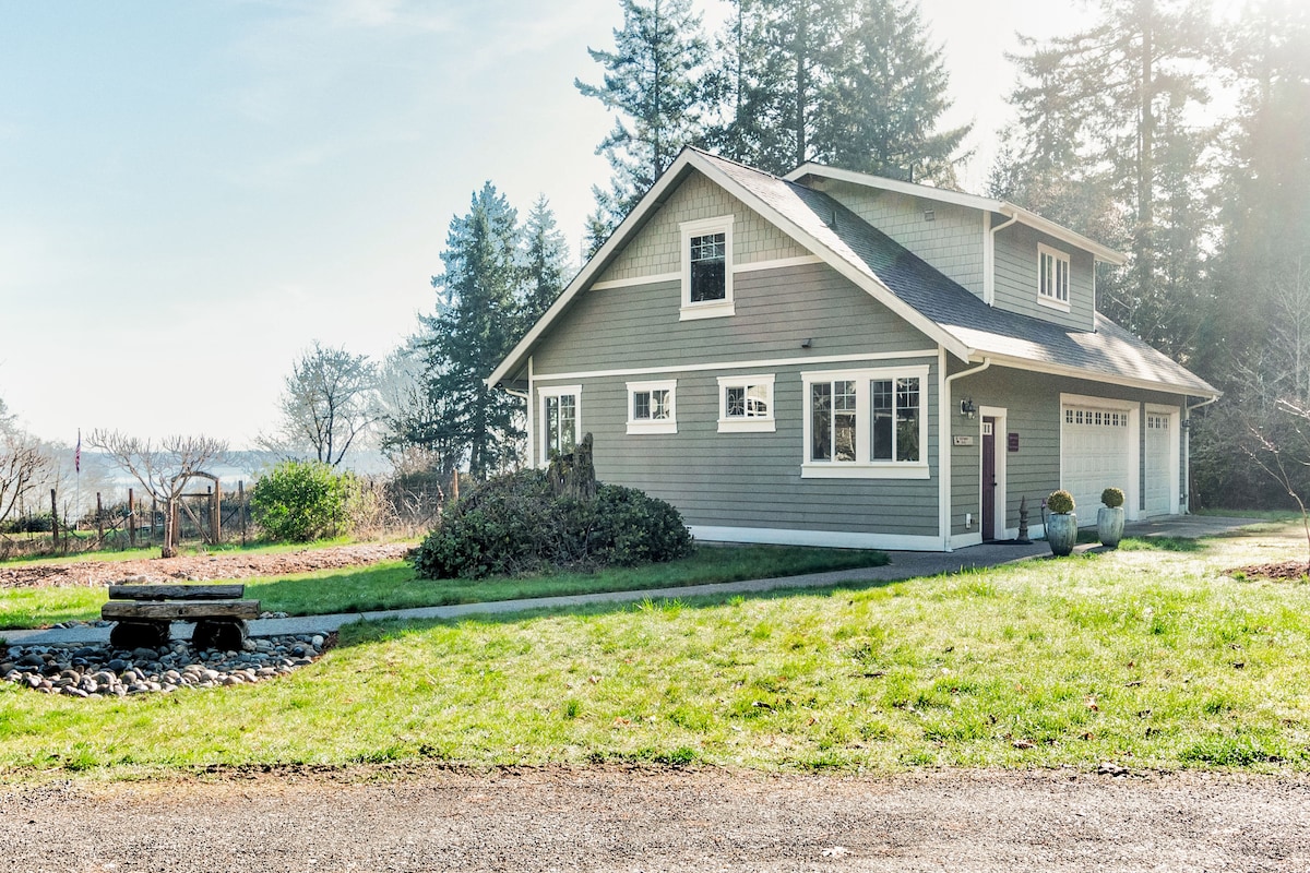 A light-colored house is positioned on a grassy area, with trees in the background. A stone fire pit can be seen in the foreground, and vineyard spaces are visible in the distance. Windows allow natural light to enter the home, creating an inviting exterior presence.