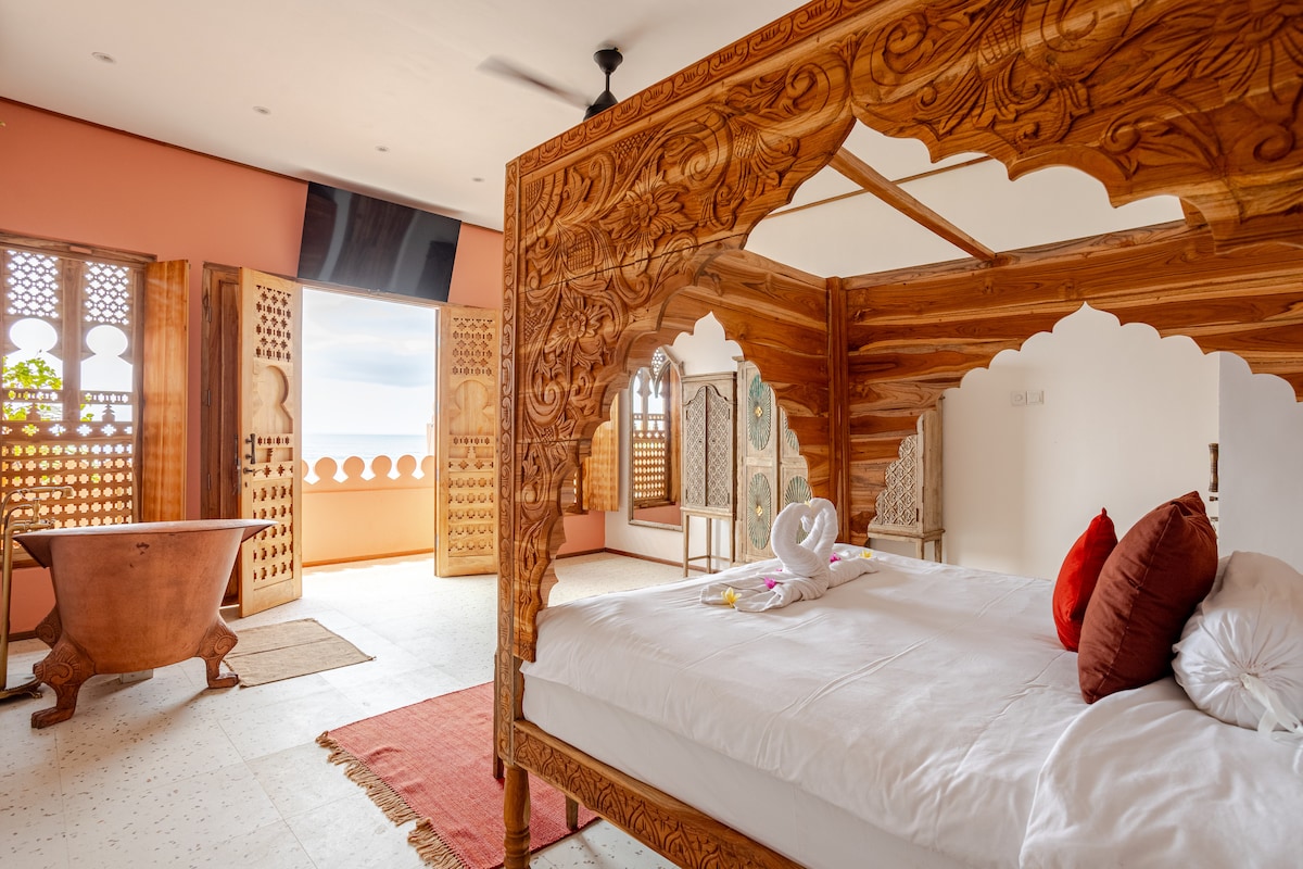 A spacious bedroom features intricate wooden carvings and a large four-poster bed. A cozy bathtub is positioned nearby, and large windows provide views of the outside. Textured rugs and elegant decor create a relaxing ambiance.