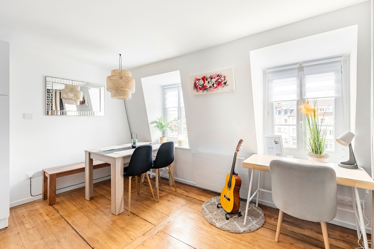 A bright dining area features a wooden table with four black chairs beside a bench. A desk with a chair is positioned near the window, accompanied by a small plant and a stylish lamp. A guitar is displayed on the floor, and natural light fills the space.