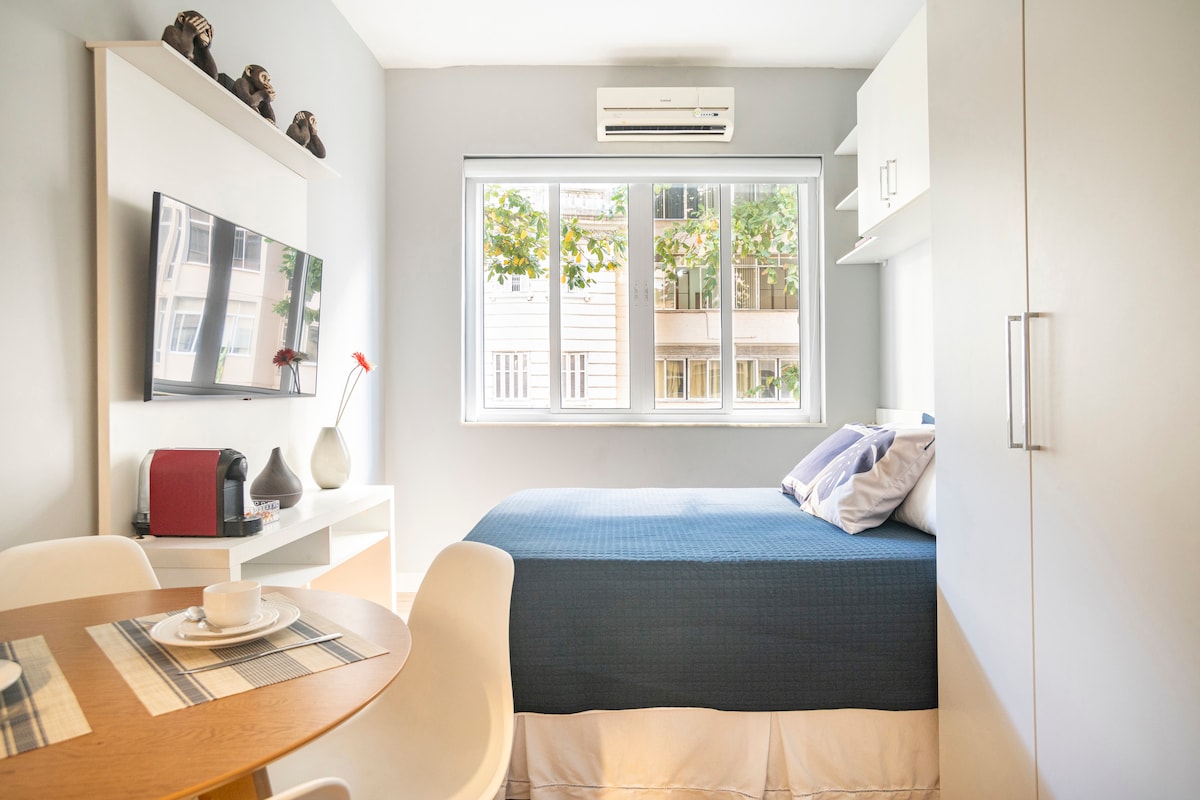 A serene studio space features a spacious bed with a blue coverlet, positioned against light grey walls. A small dining table holds a place setting, and a television is mounted on a wall opposite the bed. Natural light floods in through a large window, framed by white shutters.