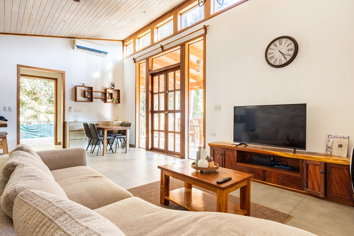 A spacious living area is highlighted by a large sectional sofa positioned beside a wooden coffee table. Natural light pours in through expansive glass doors. A television is mounted on a wooden console, with a dining area visible in the background, featuring a simple table and chairs.