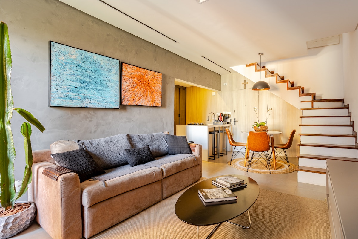 A comfortable living area displays a light-colored sofa adorned with dark cushions. Two vibrant artworks are mounted on the wall, complementing a dining space with a round table and four chairs. A staircase is visible in the background, leading to an upper level.