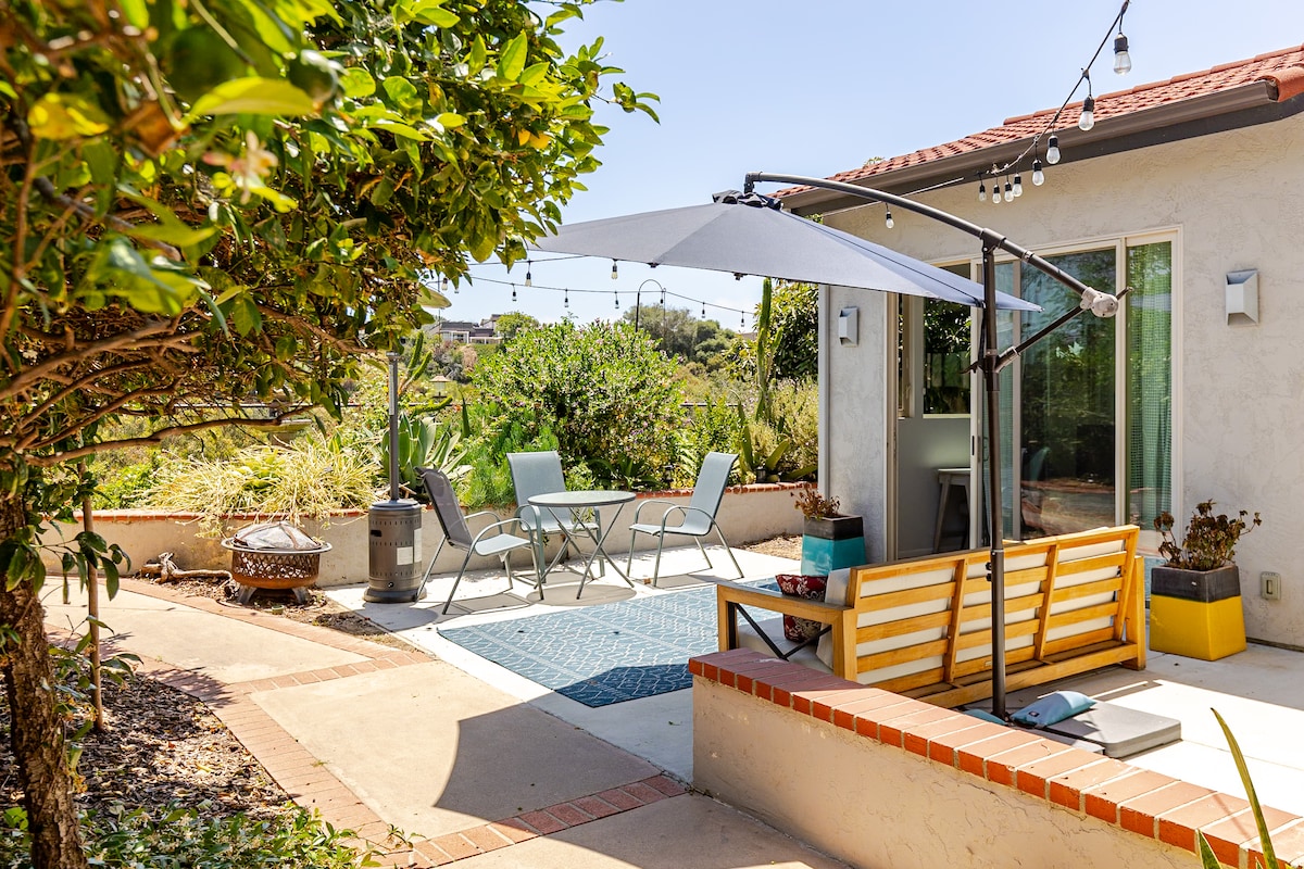An outdoor patio area is featured, showcasing a seating arrangement with a table and chairs beneath a large umbrella. Surrounding greenery and decorative lights enhance the tranquil setting. A cozy bench offers additional seating, while a glimpse of a nearby entryway is visible through glass doors.