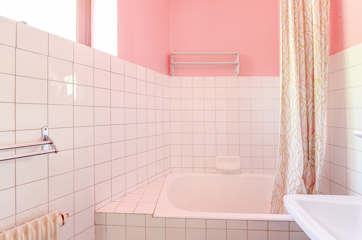 A bathroom is presented, featuring white tiled walls and a bathtub with an integrated shower. A pink accent wall adds a soft touch. Natural light enters through a window, and a decorative shower curtain adds a subtle pattern. Storage for toiletries is available on a wall-mounted shelf.