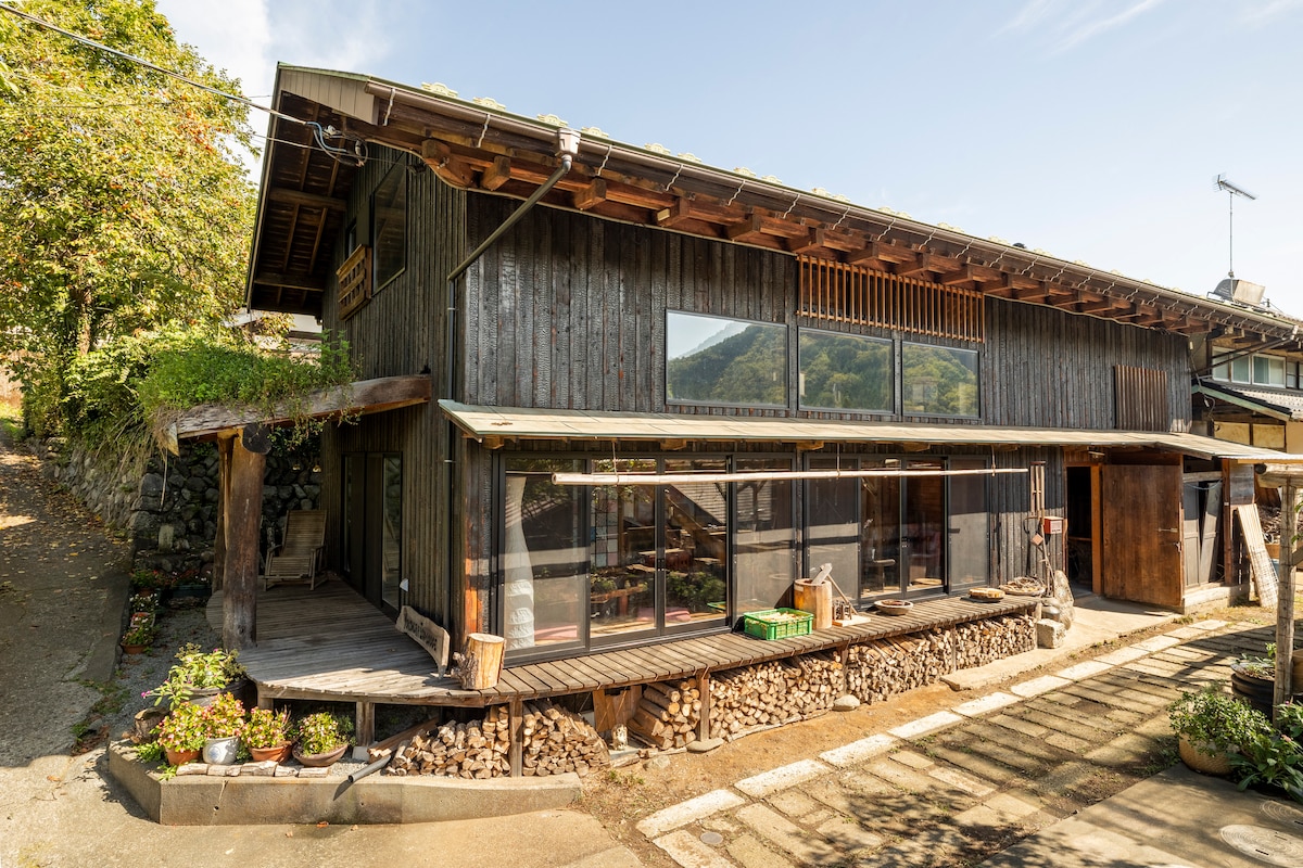The exterior of the traditional Japanese farmhouse showcases a black timber façade and large windows allowing natural light to fill the space. A wooden terrace is accentuated by stacked firewood and surrounding greenery, providing a connection to nature.
