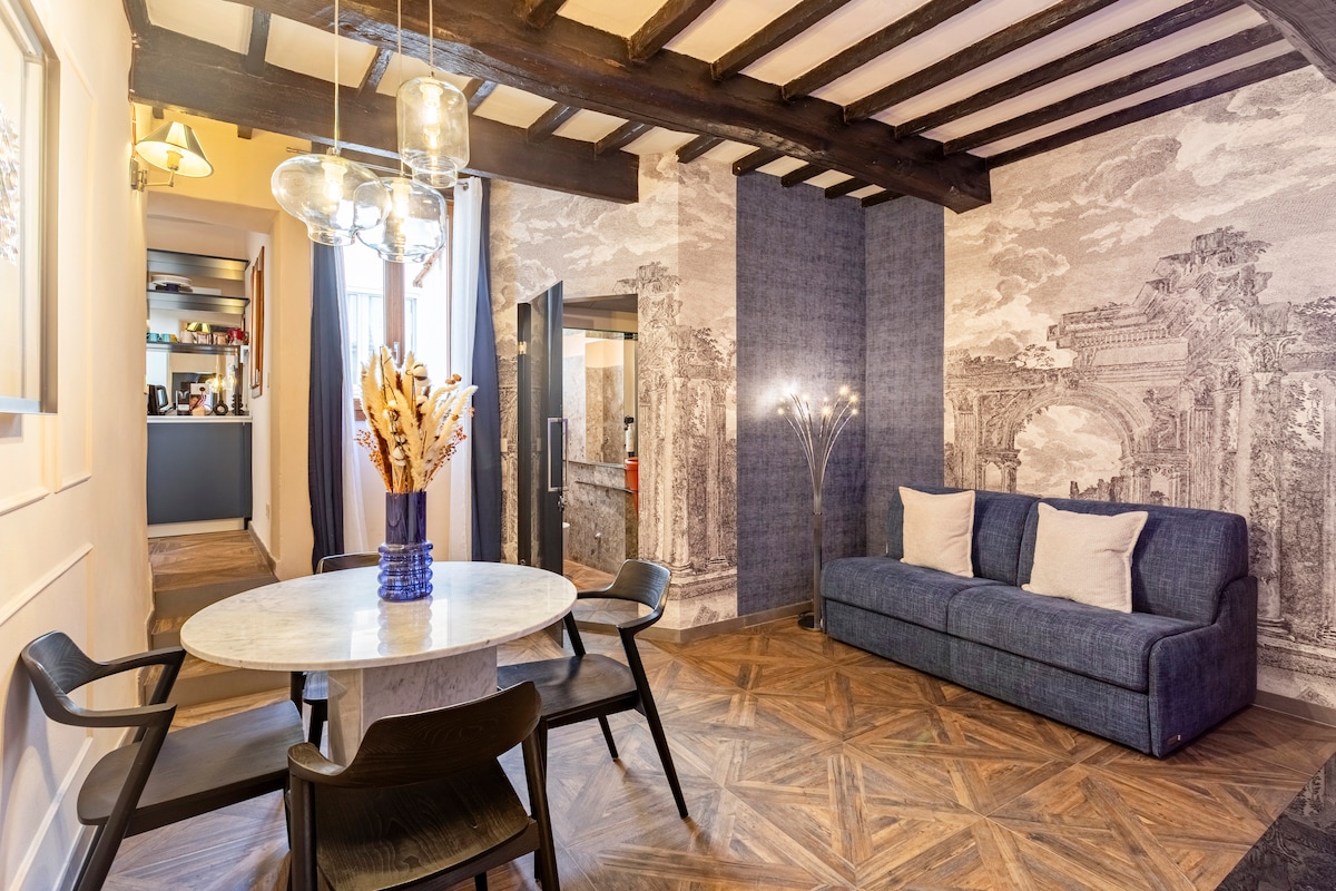 A bright living area features a round marble dining table surrounded by black chairs. A comfortable sofa is positioned against a decorative wall with intricate wallpaper. Soft lighting from a modern floor lamp complements the wooden beams above, creating a cozy ambiance.