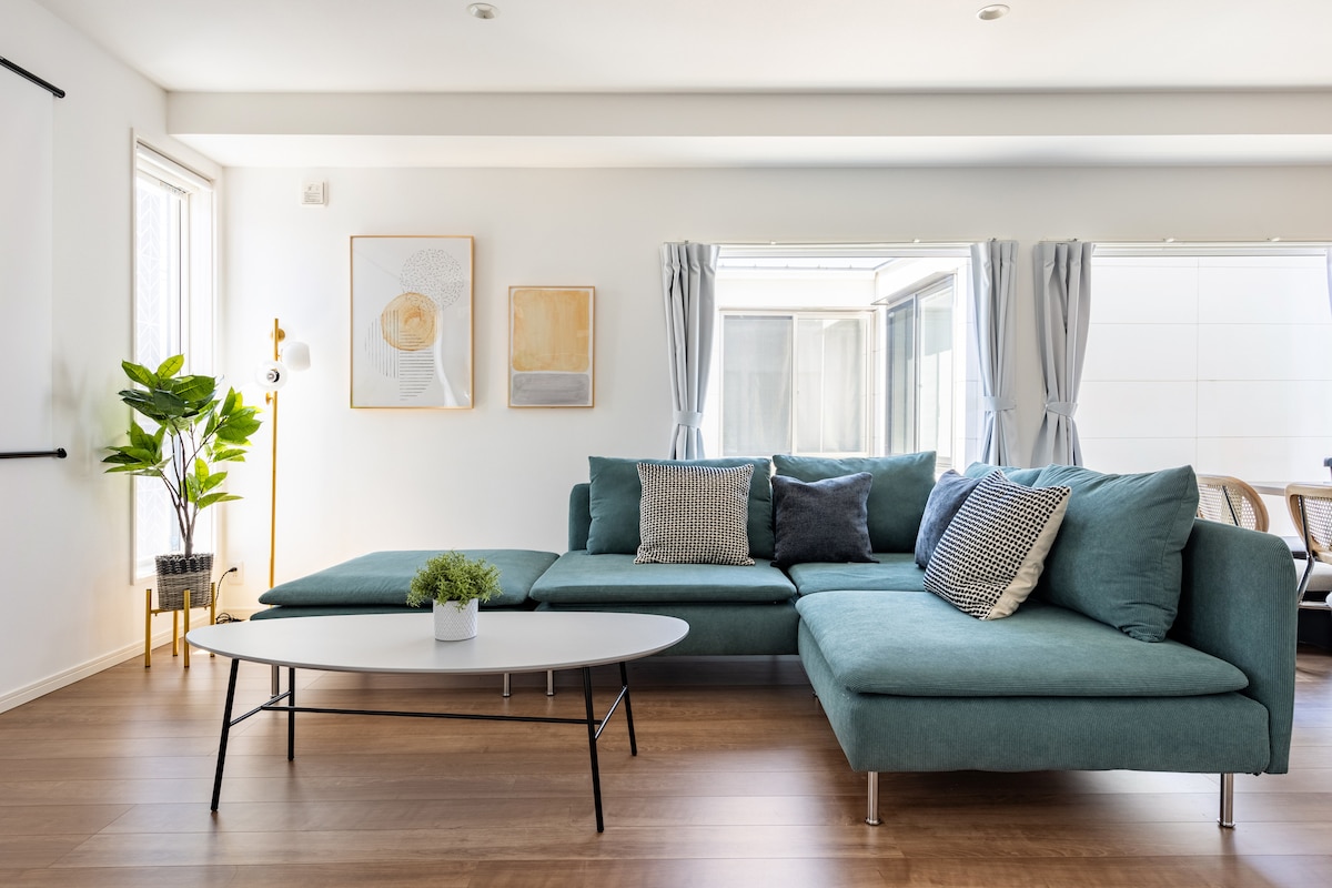 A spacious living area is filled with natural light through large windows, featuring a comfortable sectional sofa adorned with decorative cushions. A minimalist coffee table sits in front, and a potted plant adds a touch of greenery to the inviting space.