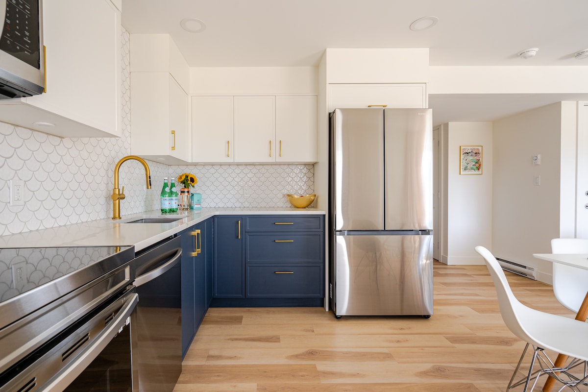 A modern kitchen features sleek white cabinetry complemented by navy-blue storage, providing ample space for cooking. Stainless steel appliances, including a refrigerator, reflect natural light. A light countertop is paired with a subtle tile backsplash, enhancing the contemporary aesthetic.