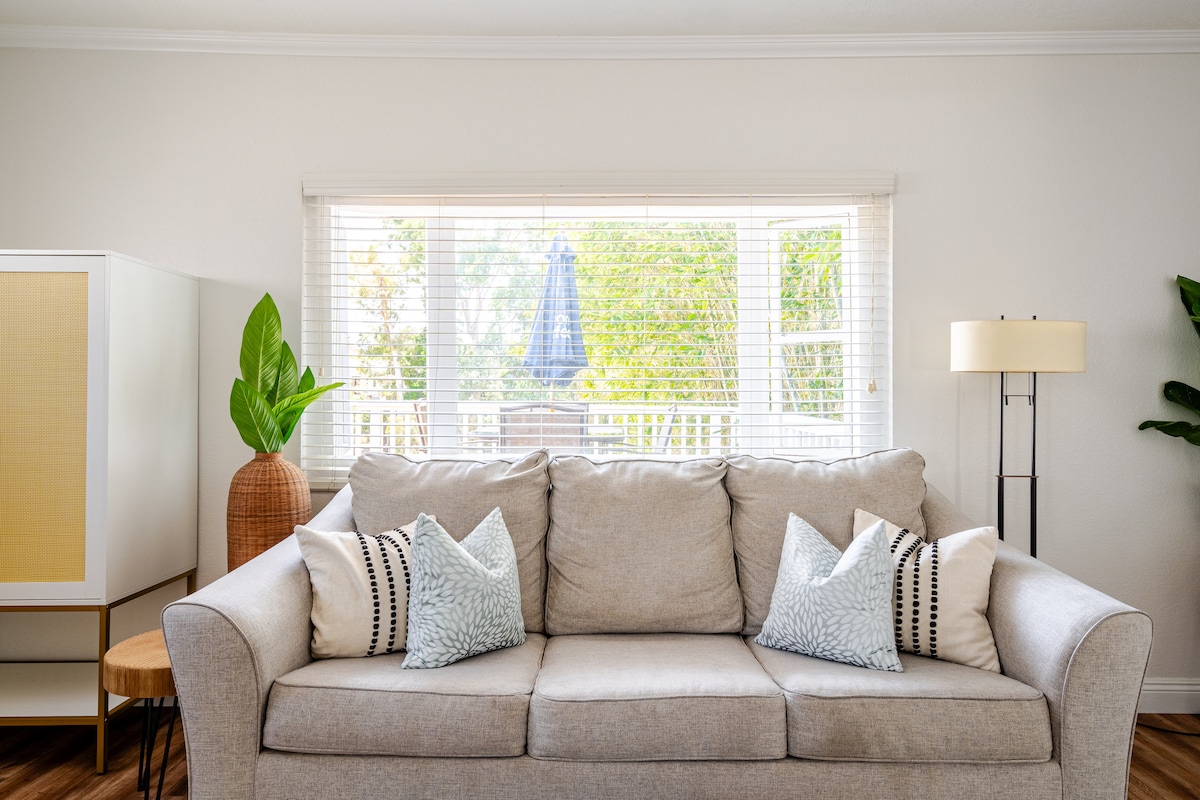 A light-filled living space features a spacious grey couch adorned with two decorative pillows. A potted plant stands nearby, while natural light filters through the window, framed by blinds that provide a view of the outdoors. A standing lamp enhances the cozy ambiance.