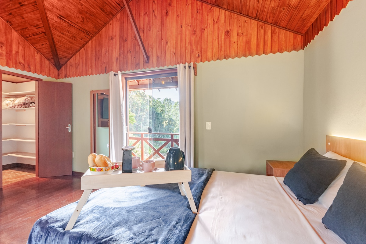 A spacious bedroom features a large bed with dark pillows and a soft blanket. A wooden tray with baked goods and a teapot is placed on the bed. Natural light enters through a window, enhancing the airy atmosphere. A closet space is visible in the background.