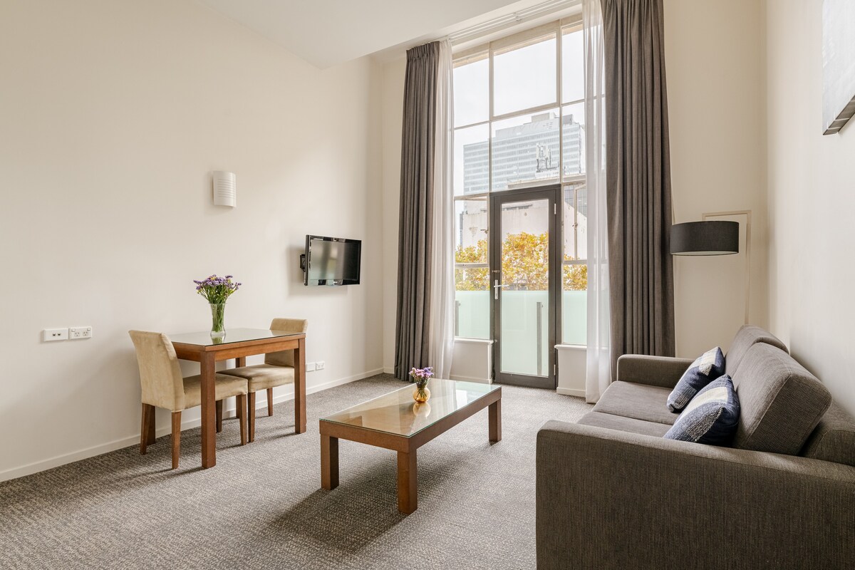 A bright living area features a cozy grey sofa and a wooden dining table with two chairs. Large windows allow natural light to fill the space, showcasing views of the city. A flat-screen TV is mounted on the wall, and neutral-toned decor complements the room.