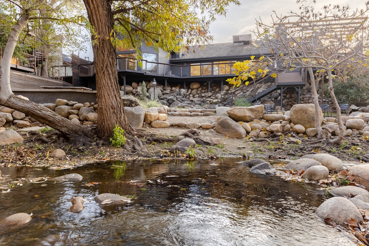 A serene riverfront scene features smooth boulders lining a gentle stream, with lush greenery around the water's edge. In the background, a modern cabin structure showcases multiple levels, surrounded by natural stones and trees, creating a harmonious connection with the landscape.
