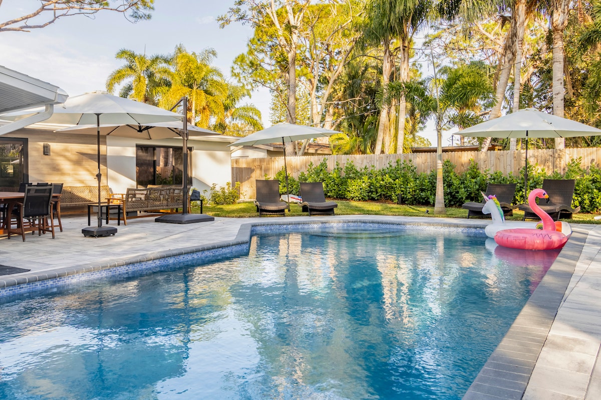 A spacious outdoor area features a large heated pool surrounded by tropical landscaping. Two shades are provided by umbrellas over four lounge chairs, while a dining table sits adjacent. Two pool floats are visible in the water, enhancing the inviting atmosphere.