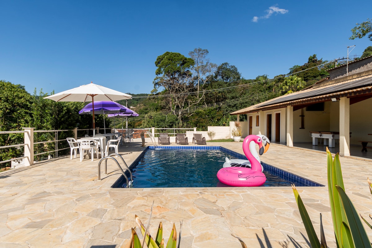 A spacious outdoor area showcases a private pool surrounded by stone tiles. A pink flamingo float is situated in the water. Umbrellas provide shade for seating arrangements, while lush greenery creates a serene backdrop.