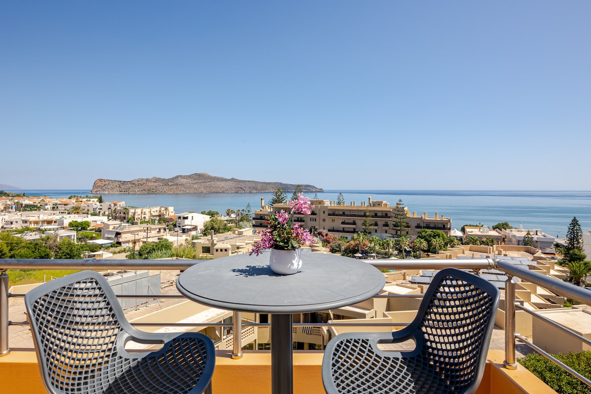 A balcony view features a round table and two chairs, surrounded by a scenic landscape. The sea and distant hills are visible, presenting a clear, unobstructed view. Sunlight brightens the space, highlighting a small potted plant at the center of the table.