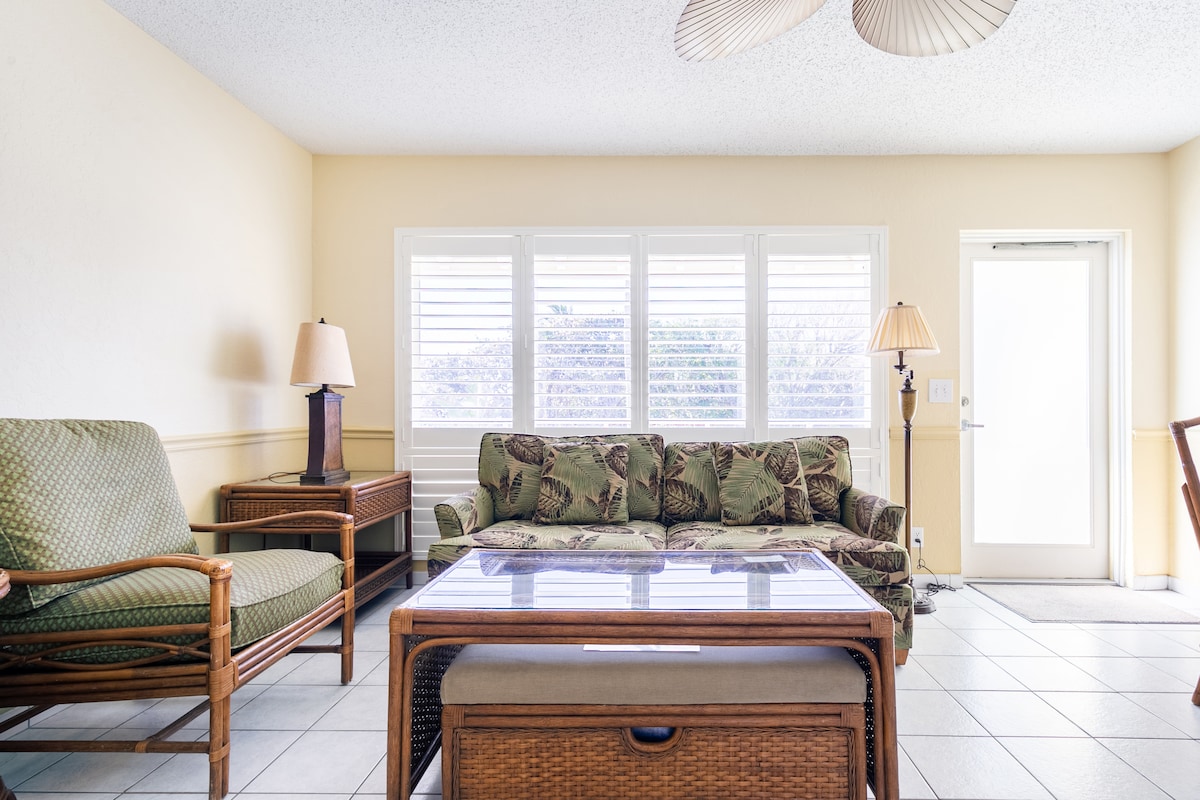 A light-filled living area features a tropical-themed sofa and a matching armchair, complemented by a glass coffee table. Natural light enters through multiple windows with plantation shutters, creating a bright and welcoming environment. A sliding door leads to an outdoor space.