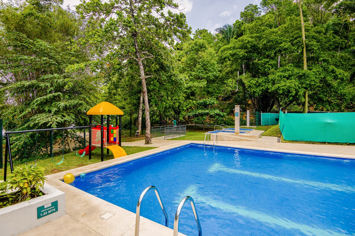 A spacious outdoor pool is surrounded by lush greenery, providing a serene environment. A children's play area with a slide and climbing structure is visible, along with lounge chairs near the water's edge, inviting relaxation in a natural setting.