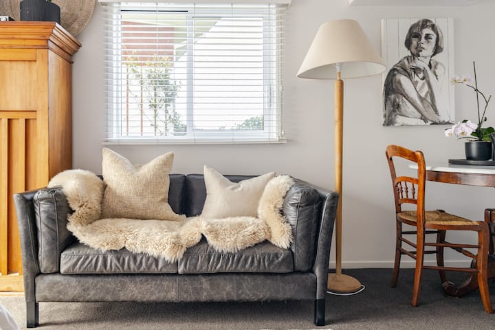 Cozy living area with a dark leather sofa adorned with soft pillows and a throw, a wooden lamp, and a small dining setup with a vintage chair