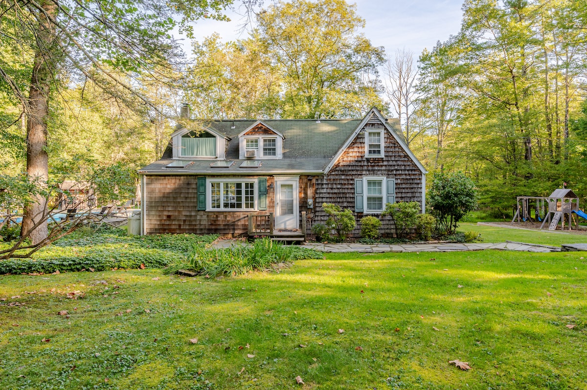 The exterior of a charming house is surrounded by lush greenery, featuring a wooden shingle facade. A welcoming front porch is visible, with steps leading to the entrance. The expansive lawn is well-maintained, and trees provide natural privacy. Play structures are seen in the background.