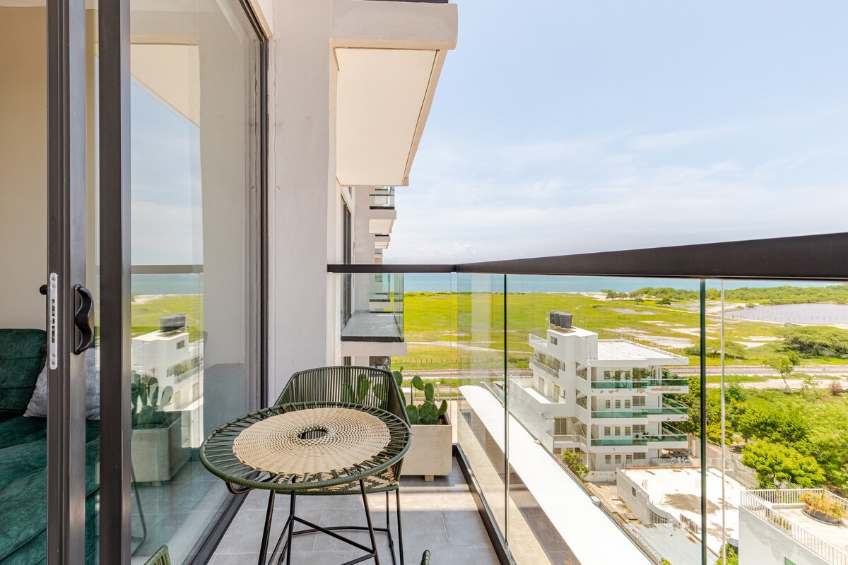 A balcony space features a small circular table accompanied by two chairs, providing a view of the surrounding landscape and water. Large glass doors frame the entrance to the interior living area, allowing natural light to fill the space.