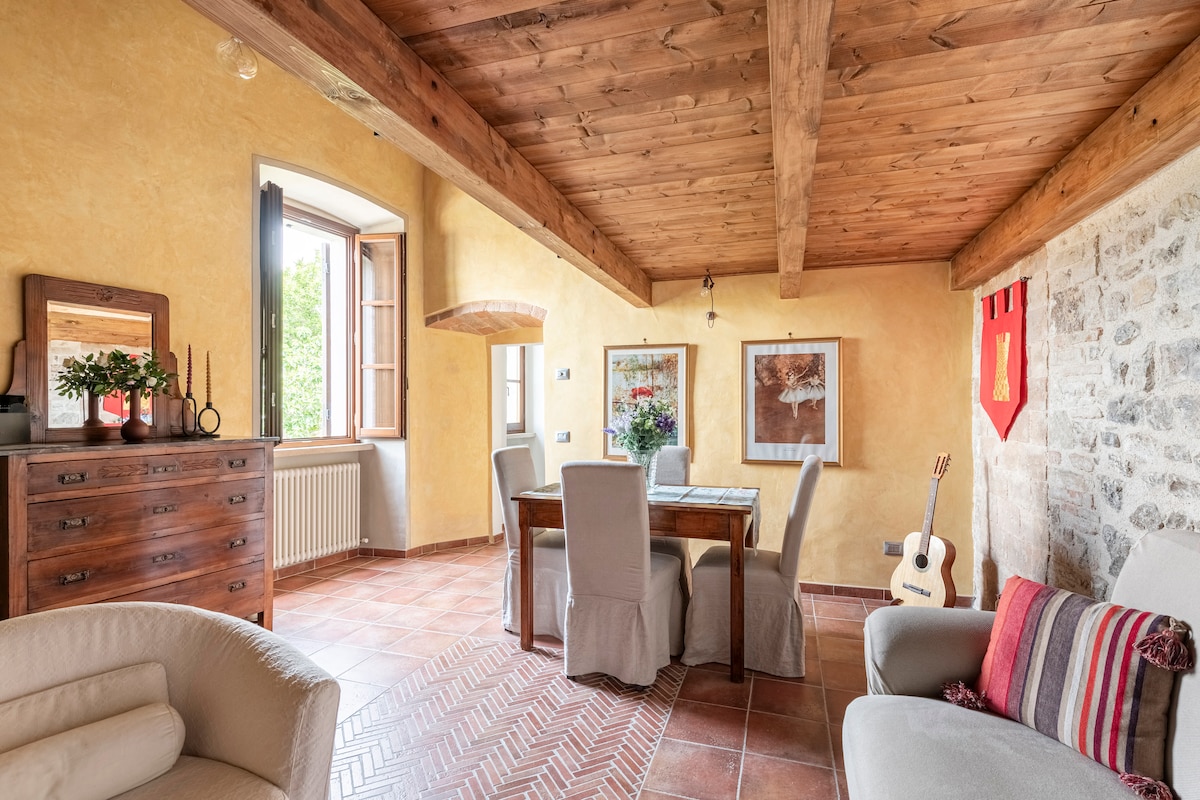 A cozy living area features a wooden beam ceiling with warm tones and terracotta flooring. A dining table with upholstered chairs is complemented by soft lighting and decorative artwork. Windows allow natural light to enter, while a guitar rests against the wall, adding a touch of character.