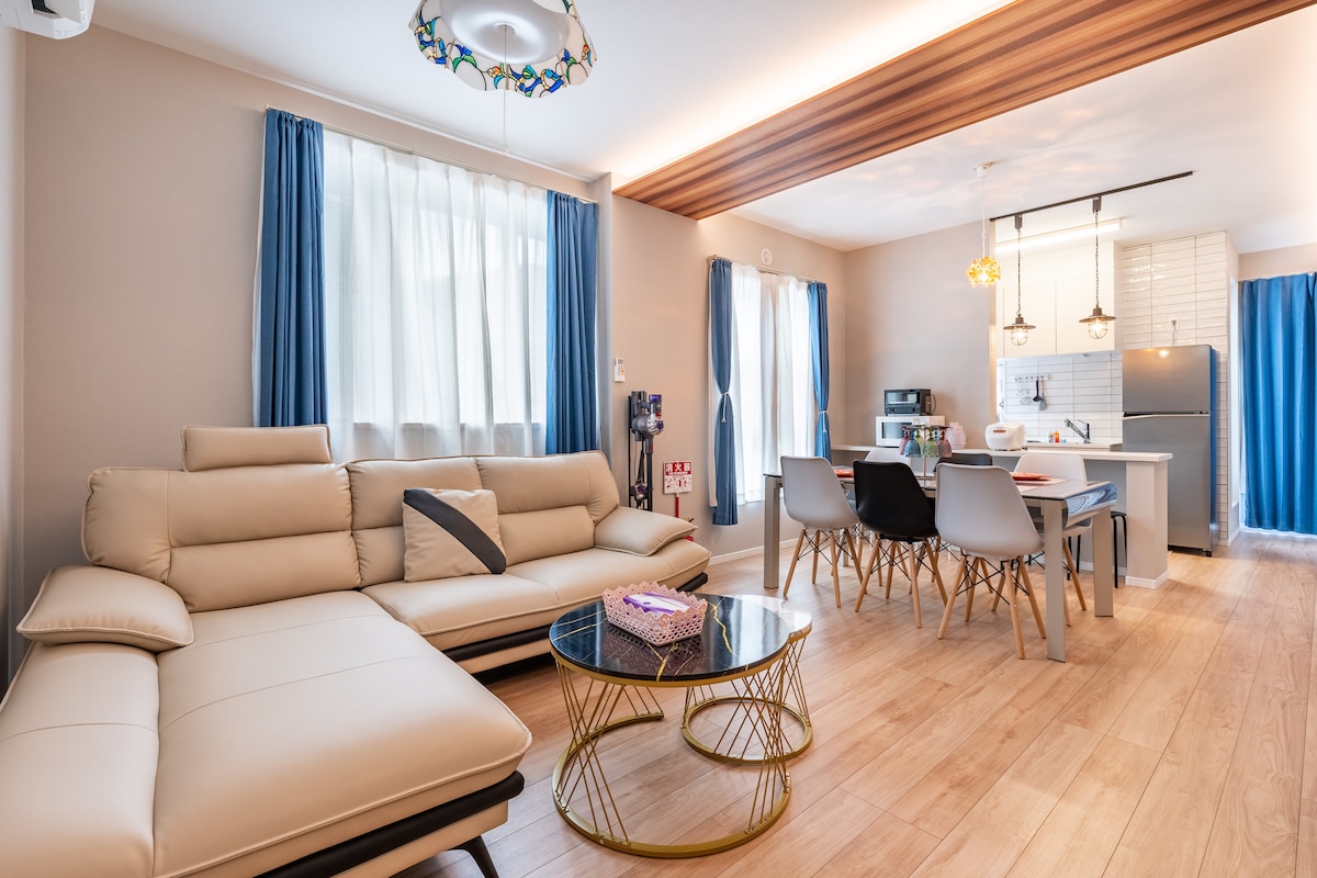 A light and airy living space is presented with a modern sectional sofa, a round coffee table, and a dining set. Large windows covered by sheer curtains allow natural light to fill the room. The adjacent kitchen features sleek appliances and a clean design.