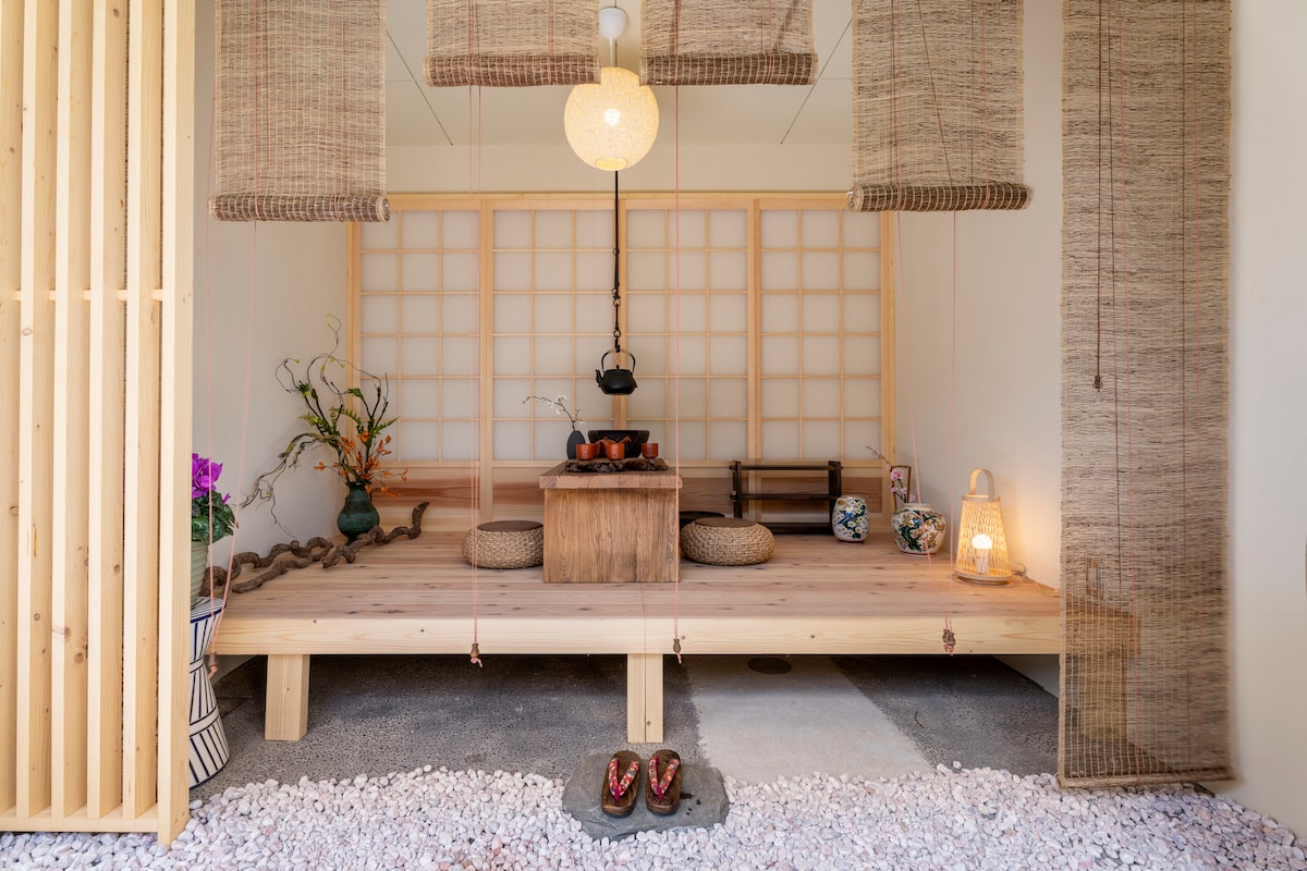 A traditional-style room features a wooden platform with seating cushions arranged around a low table. A pot hangs over a central hearth, and decorative elements include a vase with flowers and a soft light source. Natural textures and neutral tones contribute to a serene atmosphere.