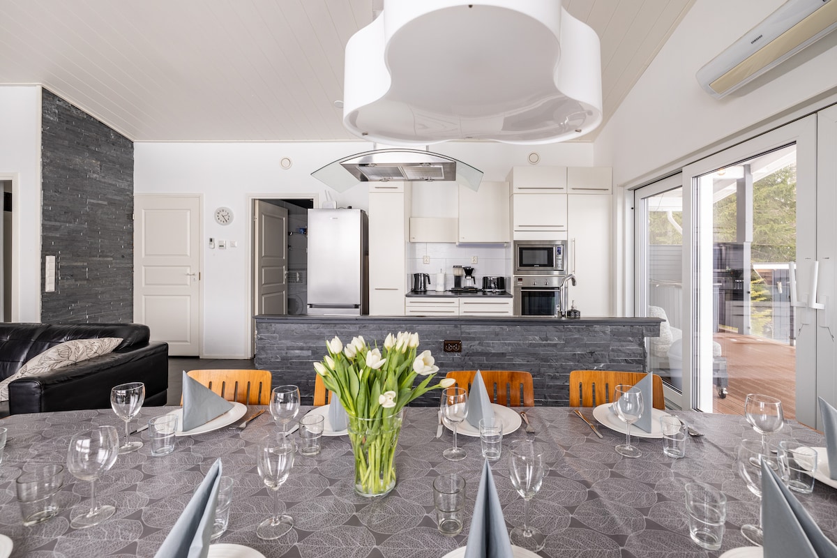 A spacious dining area features a large table set for ten, adorned with a vase of fresh flowers. Modern kitchen appliances are visible in the background, alongside a stylish kitchen island. Glass doors lead to an outdoor area, allowing natural light to fill the space.