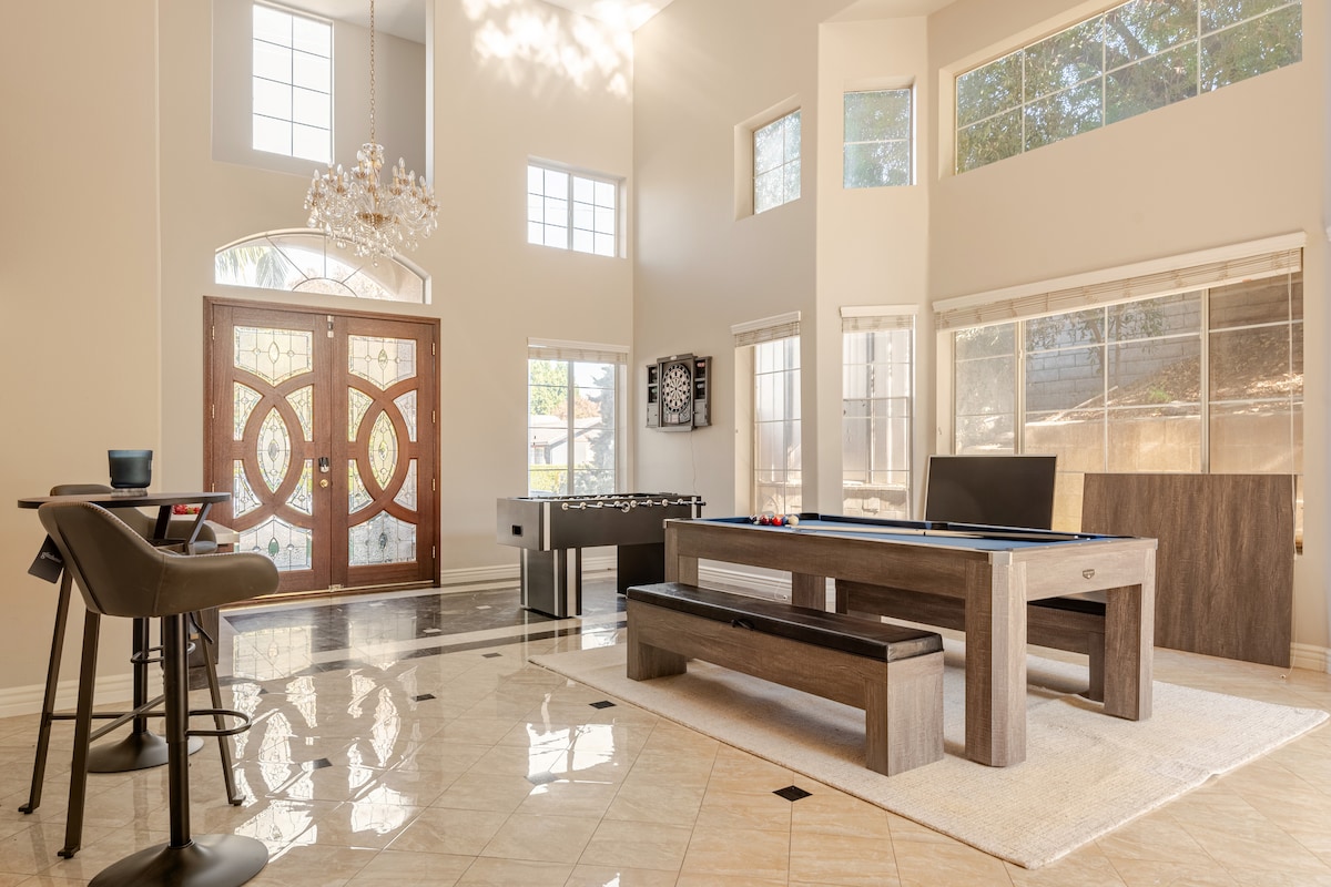 A bright and spacious game room features high ceilings and large windows that allow natural light to fill the area. A pool table and foosball table are present, complemented by a seating area with modern bar stools near an elegant entryway with ornate double doors.