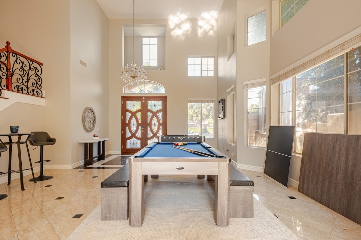 An expansive game room showcases a blue pool table at its center, surrounded by ample seating. Natural light enters through large windows, highlighting the high ceilings and elegant chandelier. A stylish entrance with decorative doors is visible in the background, enhancing the room's spacious feel.
