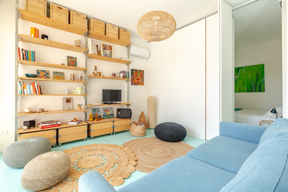 An inviting living area features an array of bookshelves with decorative items and various books. A light blue sofa is complemented by round natural fiber rugs. Soft poufs are arranged around the space, while artistic wall décor adds character. Light streams through an open doorway leading to the bedroom.
